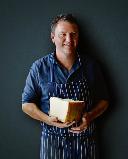 Theo Randall posant pour un portrait, regardant hors caméra, dans un tablier et tenant un gros bloc de fromage à pâte dure