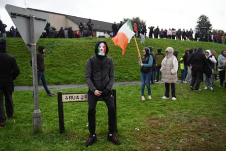Protesters, including a woman holding an Irish flag, and a young man, seated on a street sign, wearing an Irish-flag coloured balaclava under a hoodie