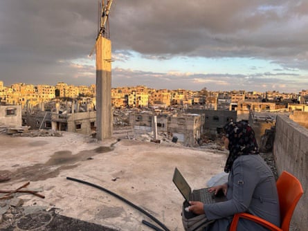 A woman sits on a plastic chair holding a laptop. She is on an exposed concrete roof, which looks out on to shells of concrete buildings and piles of rubble.