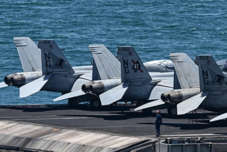 A sailor stands on an aircraft carrier’s flight deck next to several warplanes.