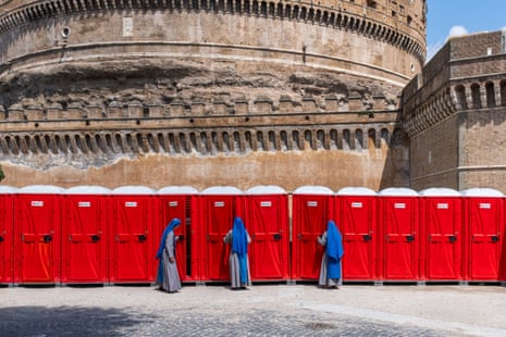 Three nuns with bright blue head coverings and grey robes peep inside the doors of bright red portable toilet cubicles in front of an ancient cylindrical stone castle.