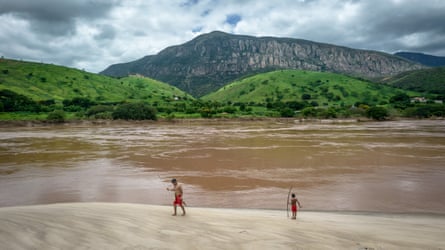 Two people walk on sand next to a large body of water, with hills behind it