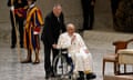 An aide pushes Pope Francis in a wheelchair as the pope makes a blessing gesture with his hand