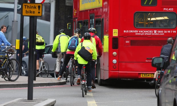 Cyclists in yellow jackets struggle past a bus turning left, next to a road sign saying “think bike!”