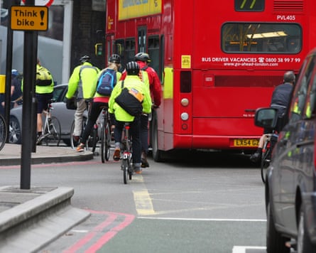 Cyclists in hi-vis jackets pedal past a red London bus on a busy road