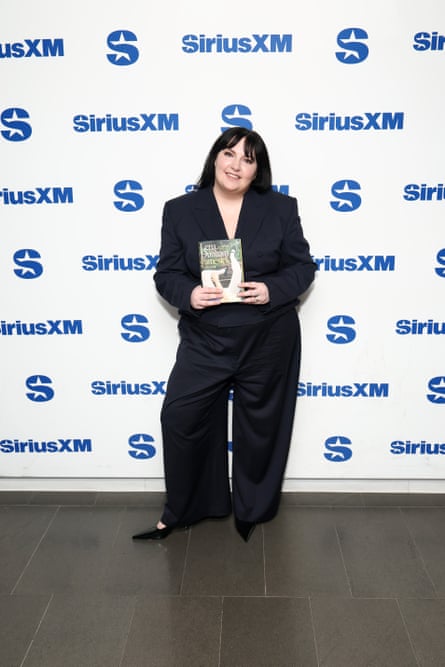Lena Dunham, wearing a black pantsuit, smiles and poses with her new memoir in front of a white backdrop dotted with the blue Sirius XM logo.