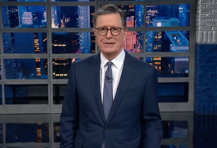 Stephen Colbert, in a navy suit, gestures while speaking in front of a city skyline backdrop