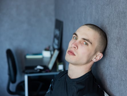 An unsmiling young man pinch cropped hairsbreadth leans against a wall
