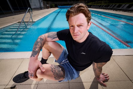 The British swimmer Adam Ramsay-Peaty, a white man wearing a black t-shirt, blue shorts and black socks, poses for the camera by the edge of a swimming pool.