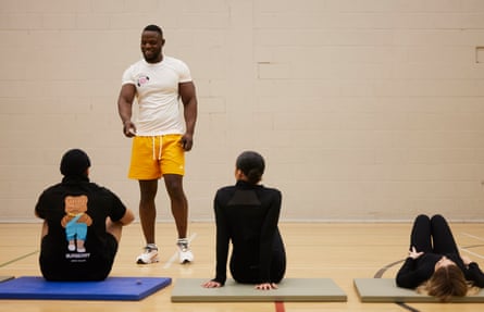 Glodi Wabelua stands in shorts and a T-shirt, while three students sit on exercise mats in front of him.