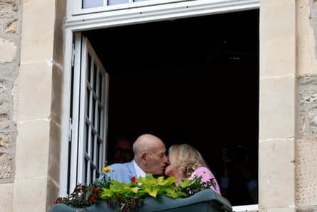 An older white couple kissing in the second-story window of a stone building with a flower box.