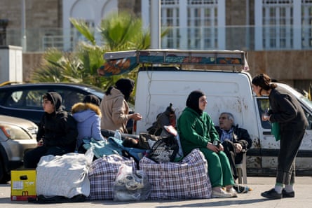 A woman talks to a family sitting on plastic chairs with large bags of belongings