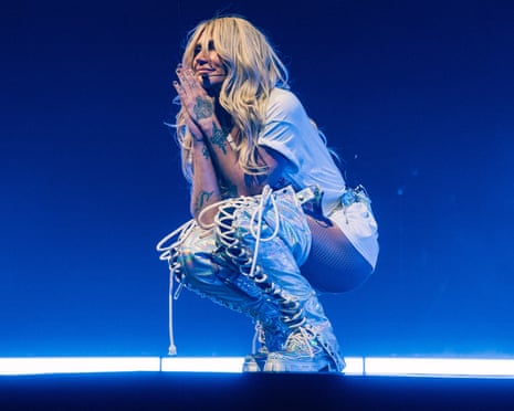 woman in knee-high boots and white T-shirt smiles, holding her hands together on stage