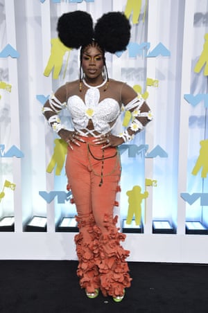 Monet X Change is posing on the black carpet. They are wearing a white top in the shape of a flower with straps around their arms, and orange trousers with ruffles around the bottoms. Their hair has been shaped into two circles which resemble mickey mouse ears