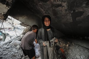 A woman with a sad expression on her face holds a dusty piece of clothing as a boy behind her gathers belongings from a collapsed building