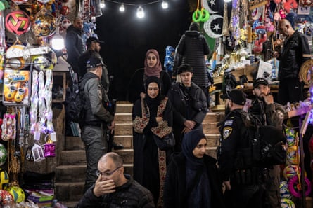 Muslim men, women and a boy walk past armed Israeli security forces as they descend steps. On each side of the steps are brightly coloured and illuminated market stalls selling toys, lanterns and festive decorations.