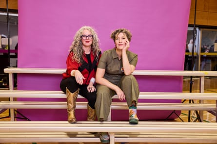 Two women sit side by side on light wooden risers with a plain magenta photo roll hanging behind them.