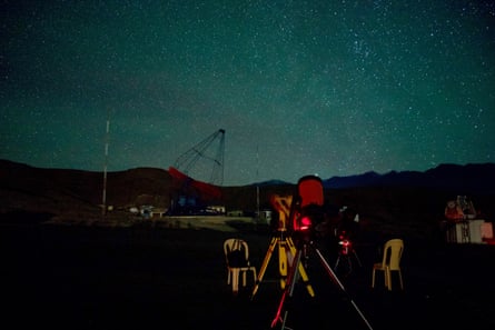 A clear night sky full of stars. In the foreground two telescopes and some plastic chairs can be made out in the darkness