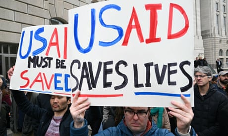 people hold signs that read 'USAID must be saved' and 'USAID saves lives'