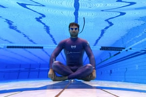 A man in a wetsuit sits cross-legged underwater near the bottom of a swimming pool