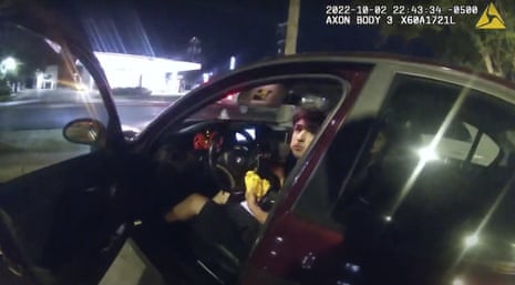 A still from police body camera video shows a teen boy sitting in a car with a hamburger in his hands looking toward the open car door.