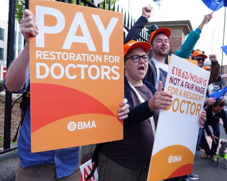Protesters hold orange signs reading 'PAY RESTORATION FOR DOCTORS' and '£18.62/HOUR IS NOT A FAIR WAGE FOR A RESIDENT DOCTOR'