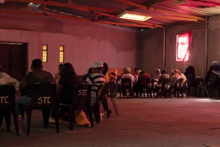 Two groups of African women sit in circles on plastic chairs in a dark hall bathed in pink light coming through cloths hung over windows