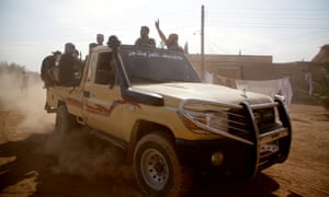 Militia holding machine guns ride in the back of a jeep along a dusty road.