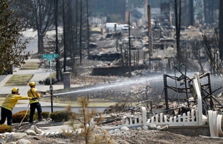 On flat, burned land, with burned trees and the ashes of what must have been houses, two people in yellow stand, one aiming a firehose.