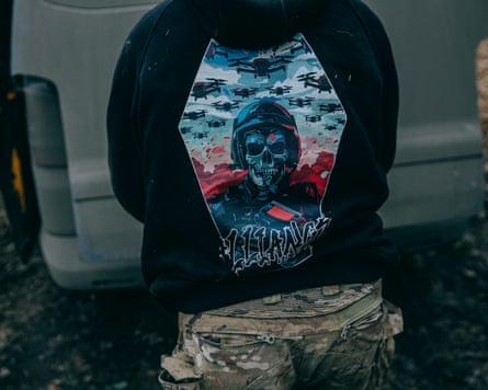A man photographed from behind wearing a black hoodie with a picture of a skull wearing a combat helmet and camouflage gear, surrounded by drones