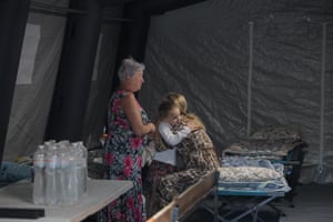 A young girl is embraced by a woman as an older woman stands by their side inside a tent with camping beds and mattresses and bottles of water on a table