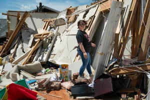 A woman inspects the remains of a roofless home, a box of Lego and a TV at her feet, the walls and doors of the home leaning diagonally in a pile