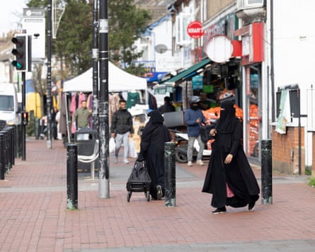 A woman with a black face covering and headscarf walks across the pavement.