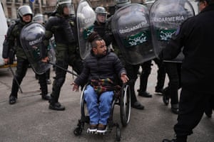 A protester in a wheelchair confronts a cordon of riot police dressed in protective gear and holding shields and batons.