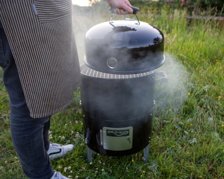 A person wearing an apron lifts the lid of the smoking Bar-Be-Quick Smoker & Grill barbecue
