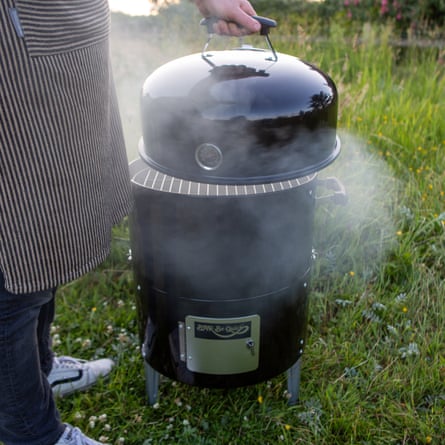 A person wearing an apron lifts the lid of the smoking Bar-Be-Quick Smoker & Grill barbecue