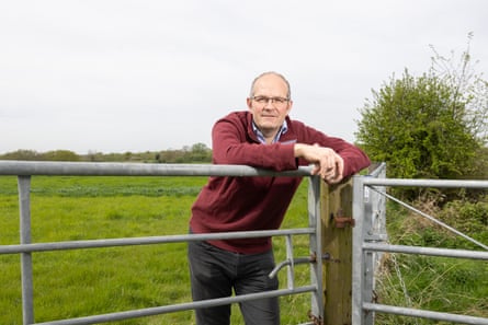 A balding man in a burgundy jumper leans on a fence at a farm