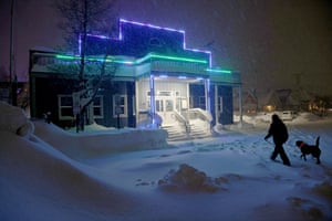 A man and his dog walk through the snow towards a building that has strips of green and purple neon lighting on it