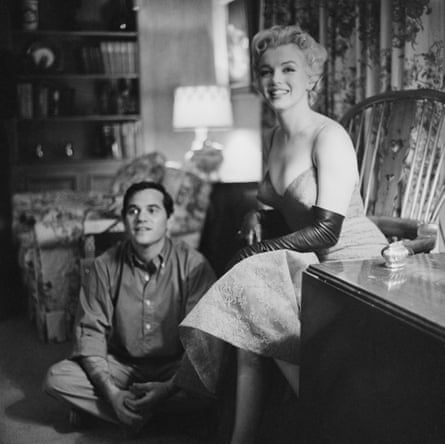 Marilyn Monroe sits in a living room with a man cross-legged on the floor by her chair