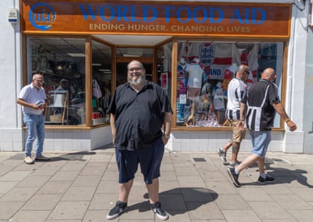 A man in a black top and dark shorts stands outside a shop smiling