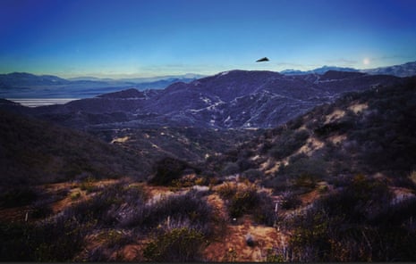 A dark triangular UFO hovers above rugged mountain ridges under a clear blue sky