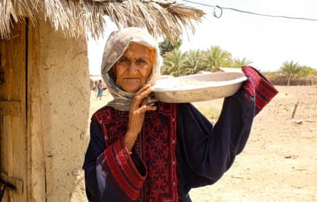 An old woman holding a plate of food outside a mud house with a straw roof.