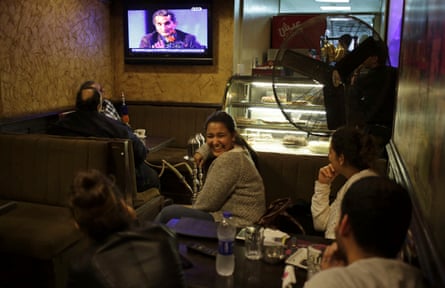 Woman wearing grey shirt sitting down at a table turns back and smiles at three other people sitting as a TV plays on in the background