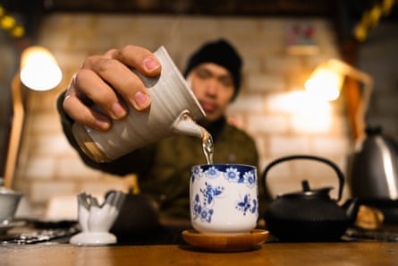 In the foreground is tea being poured from an earthenware pot into a blue and white porcelain cup. In the background is a man in a beanie.