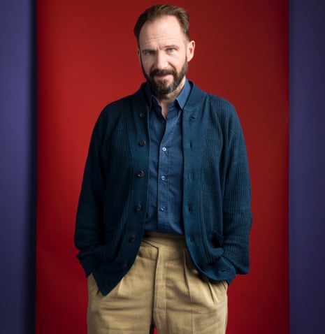 Ralph Fiennes with a beard in a shirt and cardigan, hands in his trouser pockets