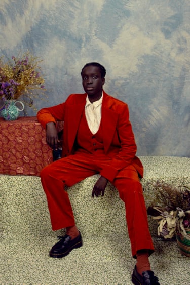 A man in a red velvet three-piece suit sits on a patterned bench beside dried flowers