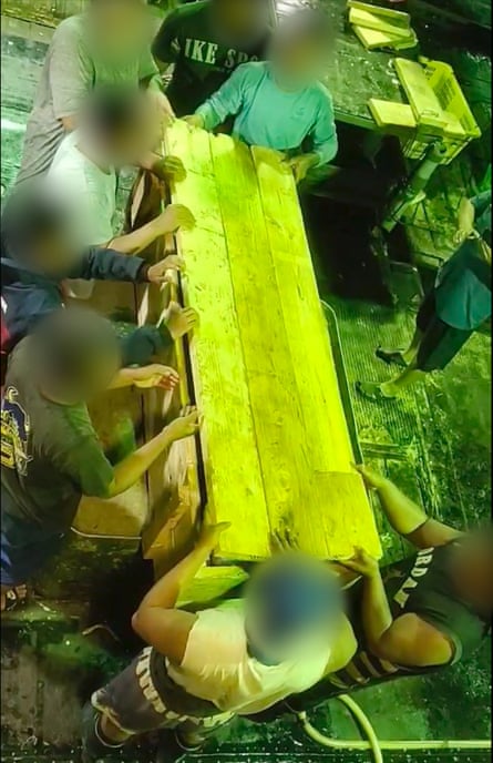 Crew members with their faces blurred stand with their hands on a wooden coffin.
