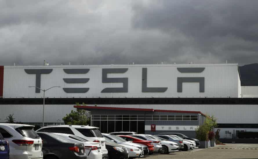 Tesla's factory in Fremont, California, a long, low white building with "Tesla" in giant letters on the front is overshadowed by ominous gray clouds.