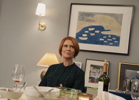 Miranda sits thoughtfully at a table beside a bottle of wine, with a picture and light in the background