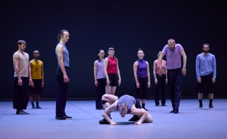 Nine people stand on stage while a tenth crawls like an animal on floor